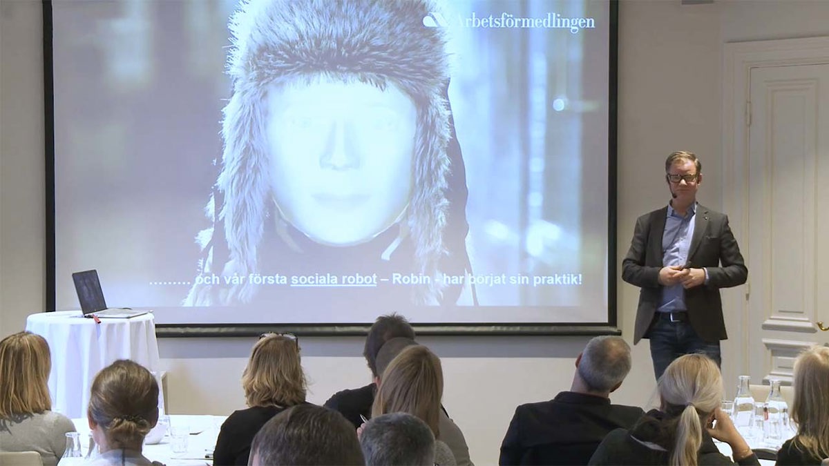 Person i föreläsarsituation stående bredvid en powerpointbild med en bild av en robot med pälsmössa.