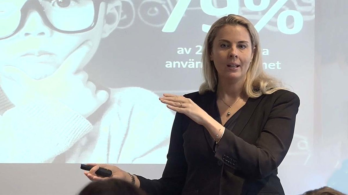 Person i seminariesituation iförd svart kavaj stående framför powertpointpresentation.