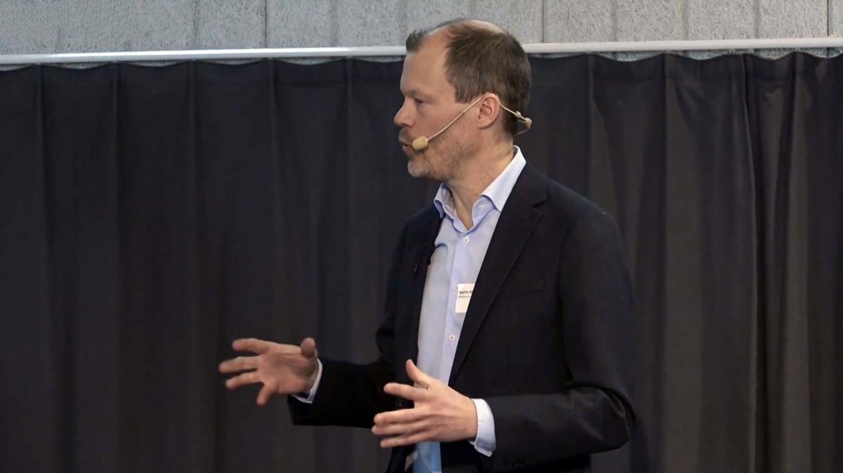 Person i föreläsningssituation iförd mikrofonheadset, svart kavaj och ljus skjorta.